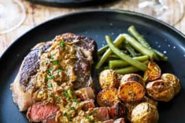 A straight-on view of a ribeye steak that has about half of it cut into sliced and is topped with a creamy gorgonzola sauce and served with roasted potatoes and green beans.