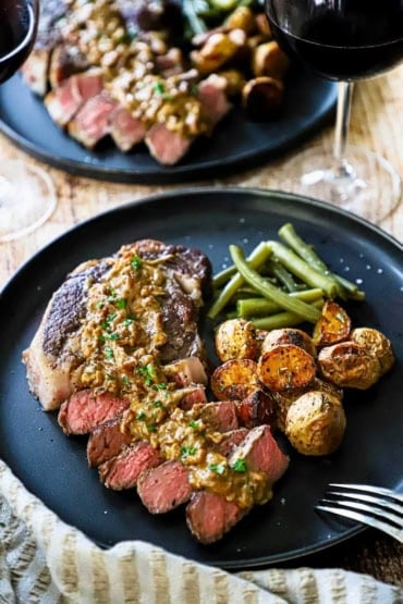 A straight-on view of a ribeye steak that has about half of it cut into sliced and is topped with a creamy gorgonzola sauce and served with roasted potatoes and green beans.