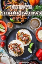 An overhead view of a round black plate filled with two open shrimp fajitas with a skilled filled with more of the fajitas nearby as well as toppings and a glass of beer.