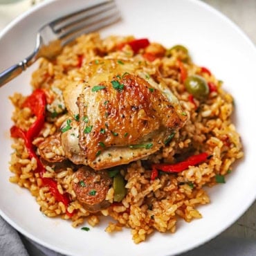 A close-up view of a serving of Spanish chicken and rice in a serving bowl with one seared chicken thigh resting on the colorful rice with slices of Spanish chorizo visible.