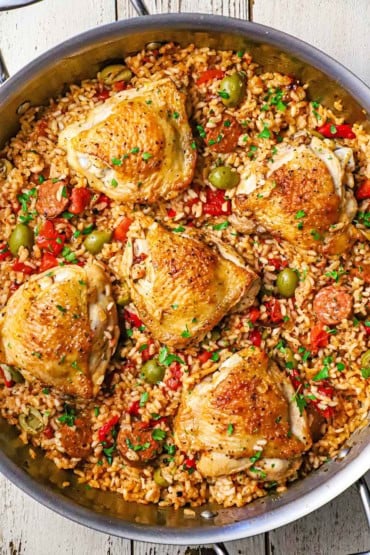 An overhead view of a large stainless steel skillet filled with Spanish chicken and rice with the seared chicken thighs resting in colorful rice along with slices of chorizo and green olives.