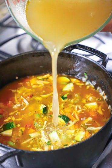 A person pouring homemade vegetable stock from a large glass measuring cup into a black pot filled with chopped zucchini, carrots, cabbage, and other vegetables.