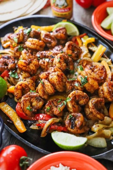 A straight-on view of a large fajita skillet filled with shrimp fajitas resting on sautéed onions and bell pepper strips with lime wedges on the side.