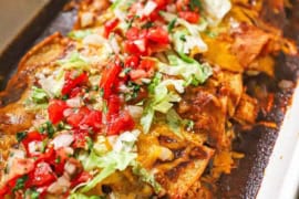 A straight-on view of a white rectangular baking dish filled with leftover turkey enchiladas with a red sauce and topped with chopped lettuce and pico de gallo.