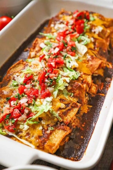 A straight-on view of a white rectangular baking dish filled with leftover turkey enchiladas with a red sauce and topped with chopped lettuce and pico de gallo.