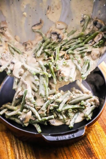 A straight-on view of cooked green beans and sautéed mushrooms in a cream sauce that are being transferred from a large skillet into a baking dish.