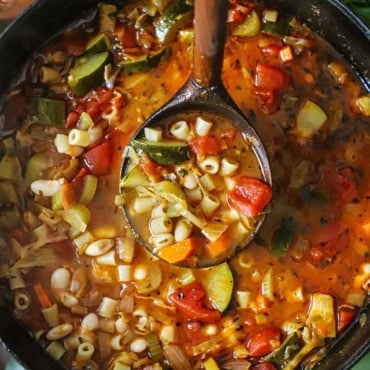 An overhead view of a black pot and a large wooden ladle lifting a serving of classic minestrone soup up out of the pot.