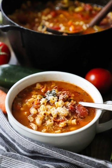 A straight-on view a white soup bowl filled with classic minestrone soup topped with grated Parmesan cheese and a pot of the soup in the background.