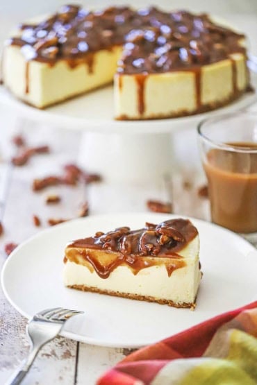 A straight-on view of a slice of pecan pie cheesecake sitting on a white dessert plate with the full cake resting on a cake stand behind the individual serving.