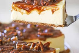 A person using a metal spatula to raise a slice of pecan pie cheesecake from the entire cake resting on a cake stand.