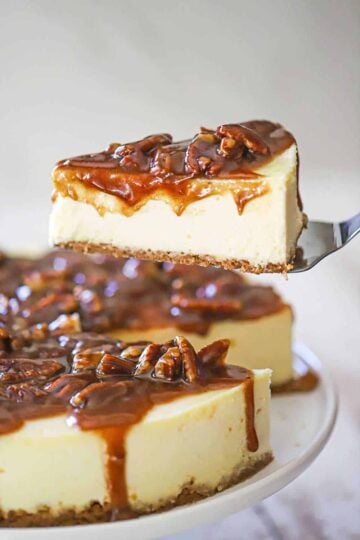 A person using a metal spatula to raise a slice of pecan pie cheesecake from the entire cake resting on a cake stand.