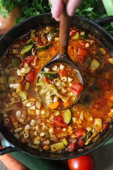 An overhead view of a black pot and a large wooden ladle lifting a serving of classic minestrone soup up out of the pot.