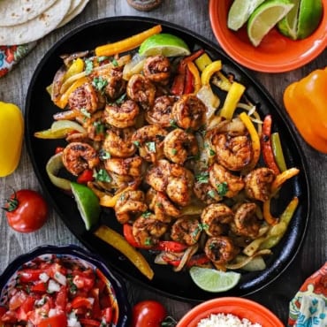 An overhead view of a large fajita skillet filled with a serving of shrimp fajitas with bowls of all the fixings nearby including pice de gallo, lime wedges, and crumbled Cotija cheese.