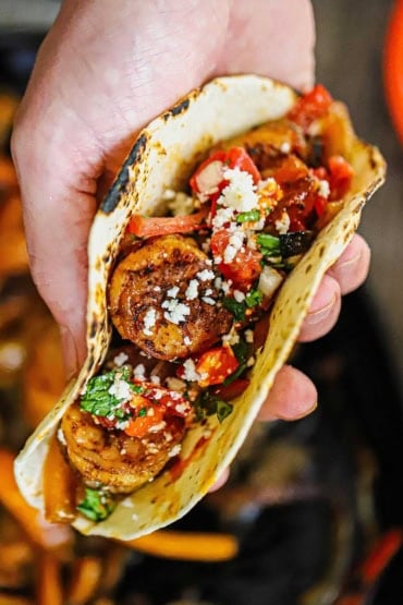 A close-up view of a person holding a prepared shrimp fajita garnished with pico de gallo, cotija cheese, and chopped fresh cilantro.