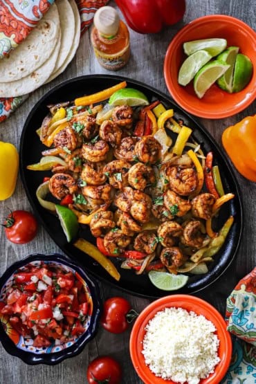 An overhead view of a large fajita skillet filled with a serving of shrimp fajitas with bowls of all the fixings nearby including pice de gallo, lime wedges, and crumbled Cotija cheese.