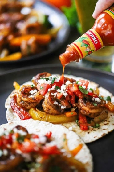 A close-up view of a person dribbling hot sauce from a bottle onto an open shrimp fajita on a lightly charred flour tortilla.