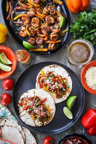 An overhead view of a round black plate filled with two open shrimp fajitas with a skilled filled with more of the fajitas nearby as well as toppings and a glass of beer.