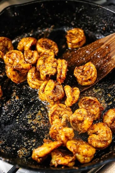 A close-up view of seasoned shrimp that are being cooked in a large black cast-iron skillet with a wooden spatula in the pan, too.