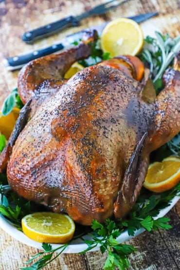 A straight-on view of a whole smoked-turkey that is resting on a white platter lined with fresh parsley and orange slices.