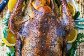 An overhead view of a whole smoked turkey that is resting on a platter of parsley and orange slices and a carving knife nearby.