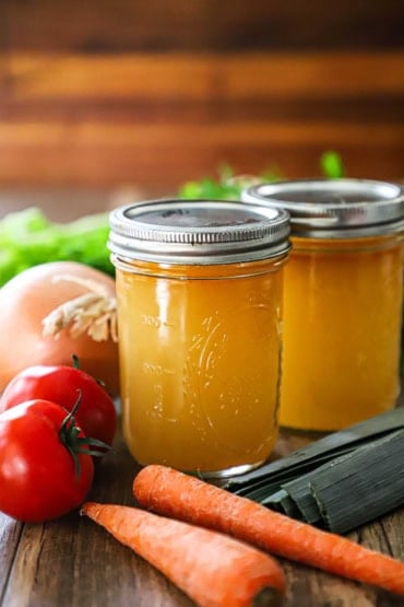 A straight-on view of two glass Mason jars that are filled with homemade vegetable stock with lids fastened on the tops of the jars and vegetables surrounding them, too.