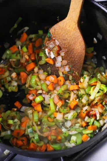 An overhead view of chopped onions, carrots, celery, and leeks being sautéed in a large black pot and being stirred with a wooden spatula.