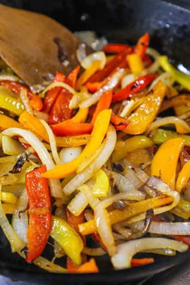 A close-up view of strips of red, yellow, and orange bell peppers and onions that are being sautéed in a large cast-iron skillet.