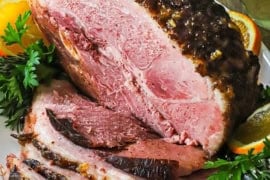 A straight-on view of a half smoked bourbon glazed ham resting on a large white platter with numerous slices cut from the roast on stacked also on the platter.