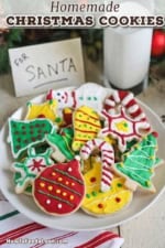 A straight-on view of a white dessert plate filled with a pile of colorful homemade Christmas cookies next to a glass of milk and a small sign that reads: For Santa.