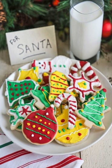 A straight-on view of a white dessert plate filled with a pile of colorful homemade Christmas cookies next to a glass of milk and a small sign that reads: For Santa.