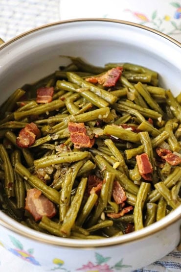 A straight-on view of a white pot that is filled with Southern-style green beans with pieces of cooked bacon intermingled.