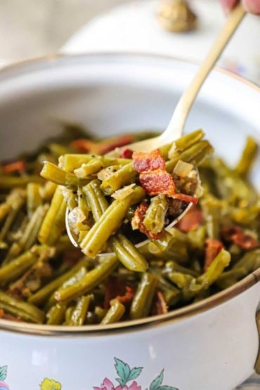 A close-up view of a person raising a large serving spoon of Southern-style green beans from an old-fashioned pot that is filled with the cooked beans.