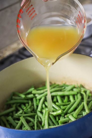 A person pouring chicken broth from a large glass measuring cup into an oval Dutch oven that is filled with fresh green beans that have been trimmed.