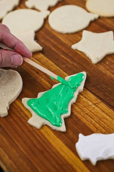 A person using a thin wooden stick to spread green royal icing across the surface of a Christmas tree-shaped sugar cookie.