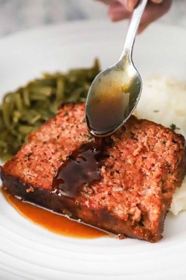 A person using a silver spoon to drizzle a glaze across the top of a glazed ham loaf slice that is resting on a bed of mashed potatoes on a white dinner plate.