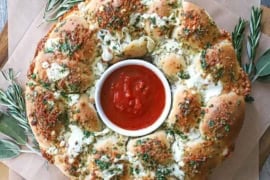 An overhead view a bread wreath with a ramekin in the center filled with marinara sauce and is surround by festive herbs.