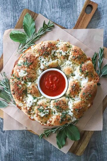 An overhead view a bread wreath with a ramekin in the center filled with marinara sauce and is surround by festive herbs.