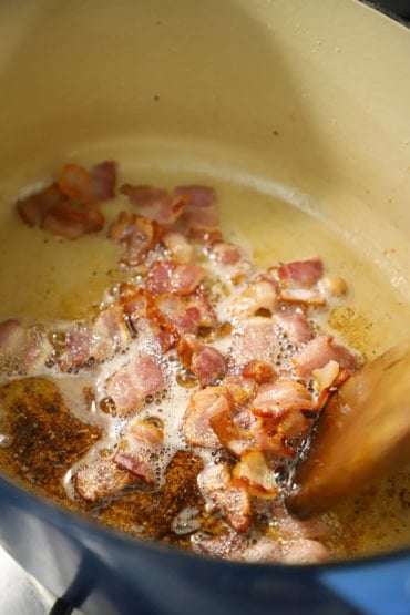 A close-up view of cut-up pieces of baking that are being fried in a large oval Dutch oven and is being stirred with a wooden spatula.