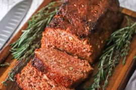 A straight-on view of a glazed ham loaf that is sitting on a small cutting board and has a few slices cut sitting next to sprigs of thyme and rosemary.