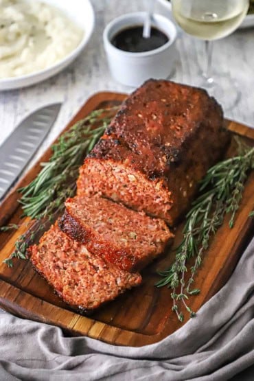 A straight-on view of a glazed ham loaf that is sitting on a small cutting board and has a few slices cut sitting next to sprigs of thyme and rosemary.