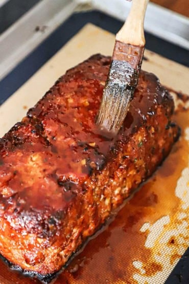 A person using a pasty brush to apply a sugar and cider vinegar glaze over the top of fully cooked glazed ham loaf.