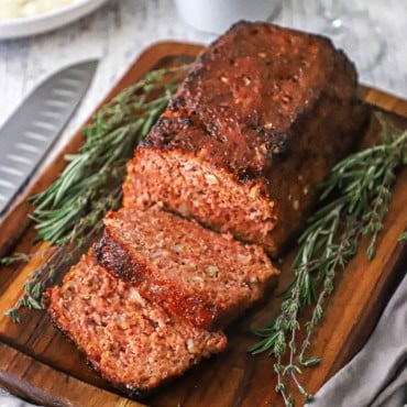 A straight-on view of a glazed ham loaf that is sitting on a small cutting board and has a few slices cut sitting next to sprigs of thyme and rosemary.