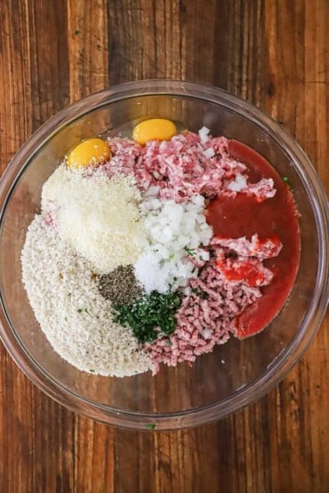 An overhead view of a glass bowl filled with ground ham, pork, breadcrumbs, eggs, chopped onion, and tomato sauce.