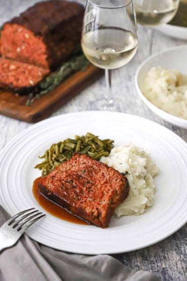 A straight-on view of a slice of glazed ham loaf that is resting on a mashed potatoes next to green beans all on a white dinner plate next to a glass of white wine.