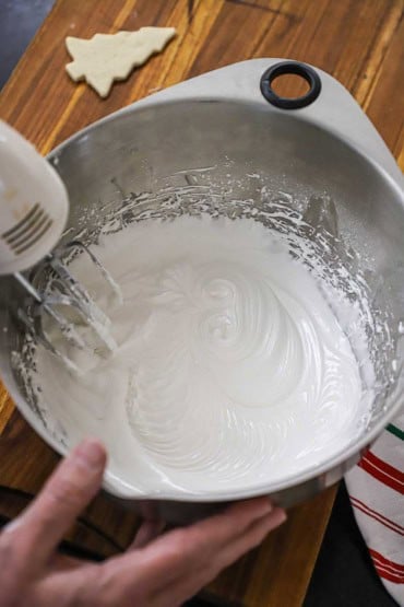 An overhead view of a person using a hand mixer to thicken white royal icing in a large stainless steel bowl.