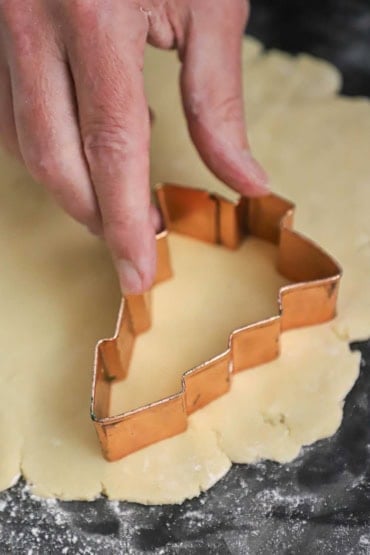 A person pressing a cookie cutter in the shape of a Christmas tree into rolled out sugar cookie dough.