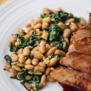 A close-up view of sautéed white beans and spinach on a white dinner plate resting next to sliced pork tenderloin.