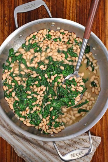 An overhead view of a large silver skillet filled with sautéed white beans and spinach with a large serving spoon in the middle of the skillet.