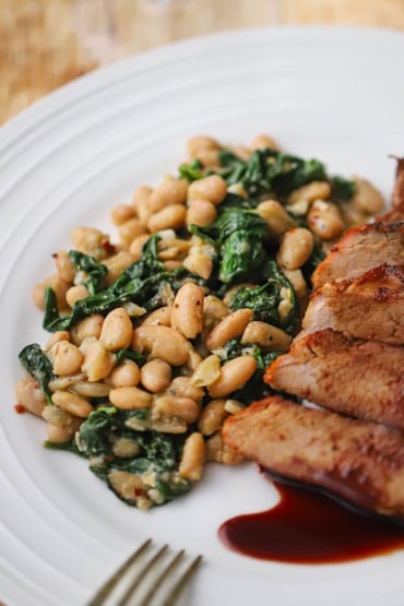 A close-up view of sautéed white beans and spinach on a white dinner plate resting next to sliced pork tenderloin.