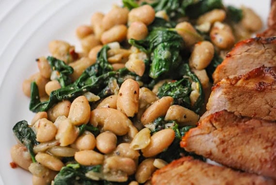 A close-up view of sautéed white beans and spinach on a white dinner plate resting next to sliced pork tenderloin.